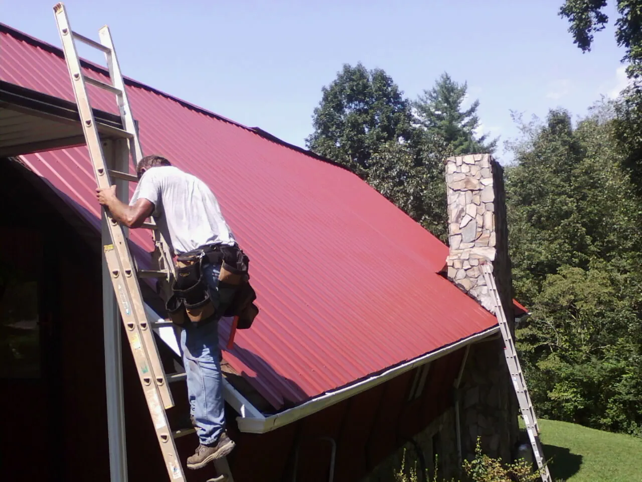 Roofer on a ladder beside a red metal roof and stone chimney — Soffit & Fascia Repair by Roofing Brothers in Port Tobacco